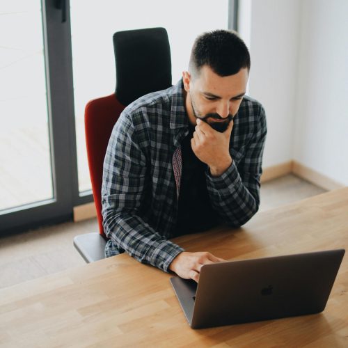 Young man with beard working on laptop at desk in a modern office setting.
